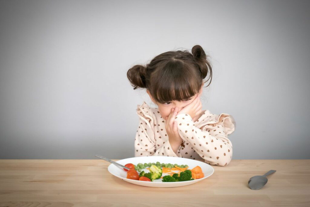 A little girl upset in front of a plate of vegetables.
