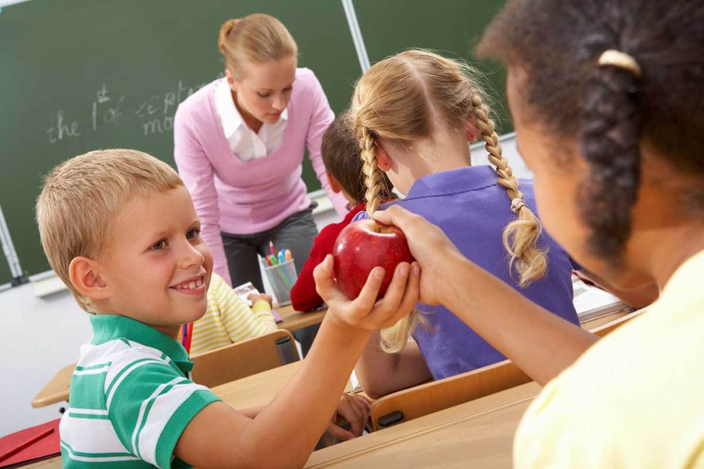 A boy handing a girl an apple in a classroom.