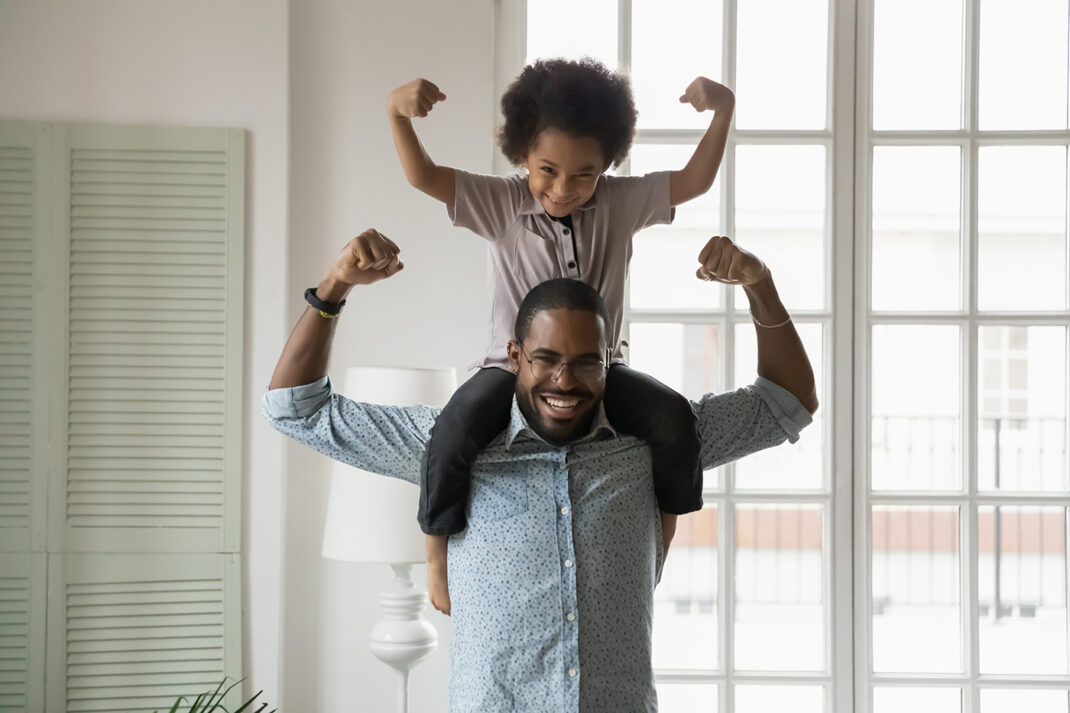A child on his father's shoulders while they both flex their arms.