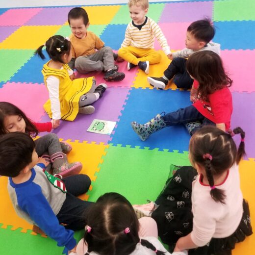 A group of children sitting in a circle holding hands on a colorful foam mat.